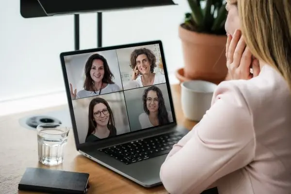 A woman sits in front of a laptop looking at a screen split between four video feeds showing smiling women.