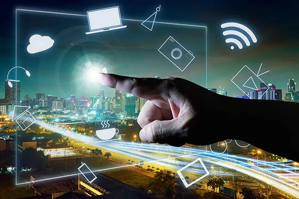 A hand touches a transparent interface containing symbols of everyday technology such as a camera, a house, and headphones; there is a modern city in the background.