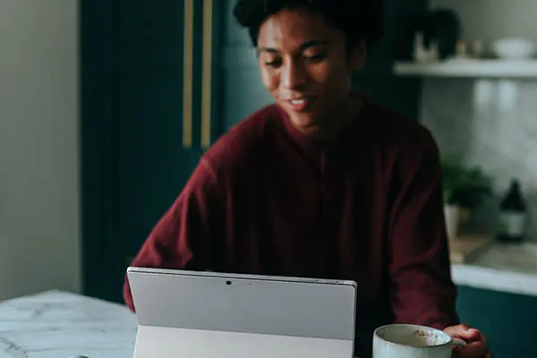 A young person drinking coffee and smiling as they look at their tablet computer.