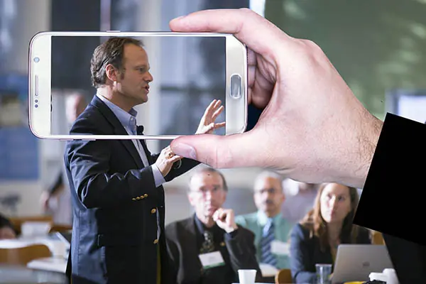 A hand holding up a camera phone to frame a speaker at a meeting.