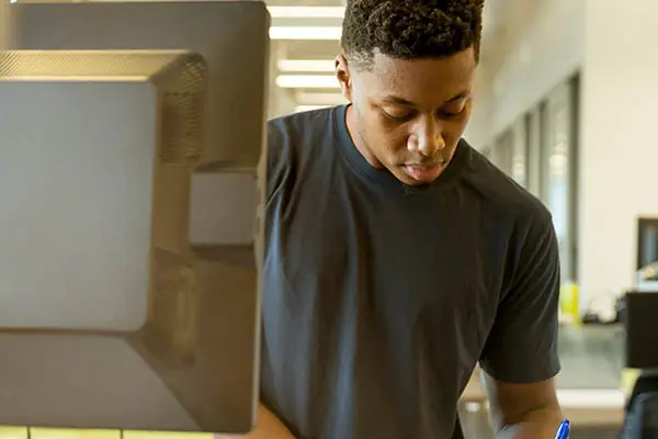 A young man makes notes while standing in front of a monitor.