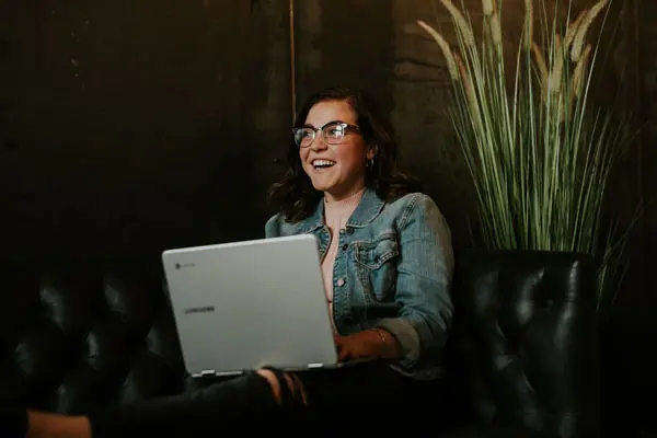 A smiling woman sits on a couch with a laptop on her knees.