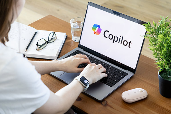 Person typing on a laptop displaying the Copilot logo, with a notebook, glasses, and a plant on the desk.