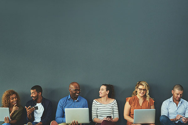 A group of six people sitting in a row against a plain grey wall, using laptops and mobile devices